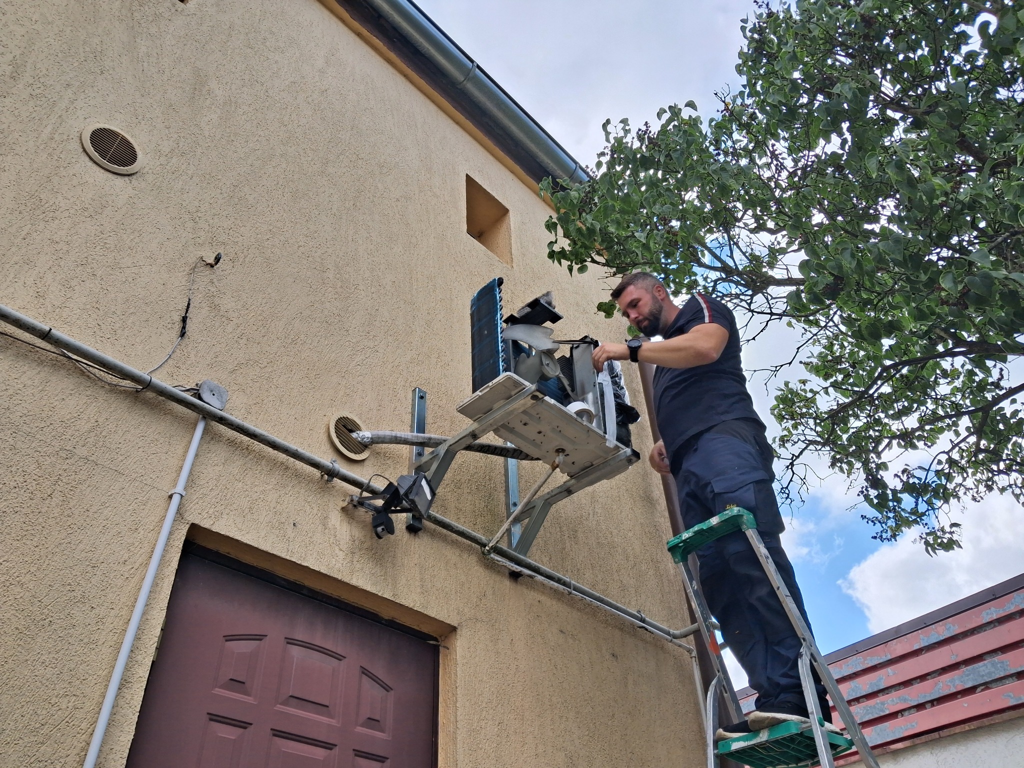 Mężczyzna na drabinie serwisuje klimatyzator na elewacji budynku, widoczne rury i elementy instalacji, drzewo w tle.