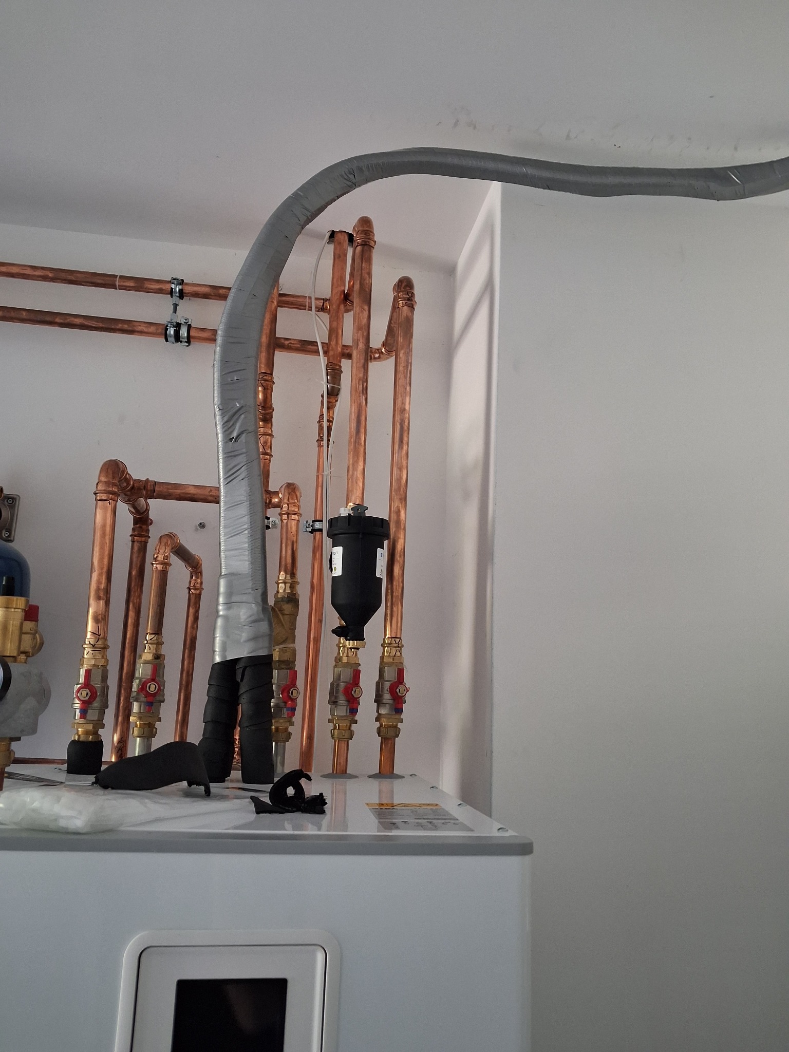 Instalacja z miedzianych rur i szarego przewodu, prawdopodobnie wentylacyjnego, zamontowana na białym urządzeniu. Widoczne zawory i elementy montażowe.