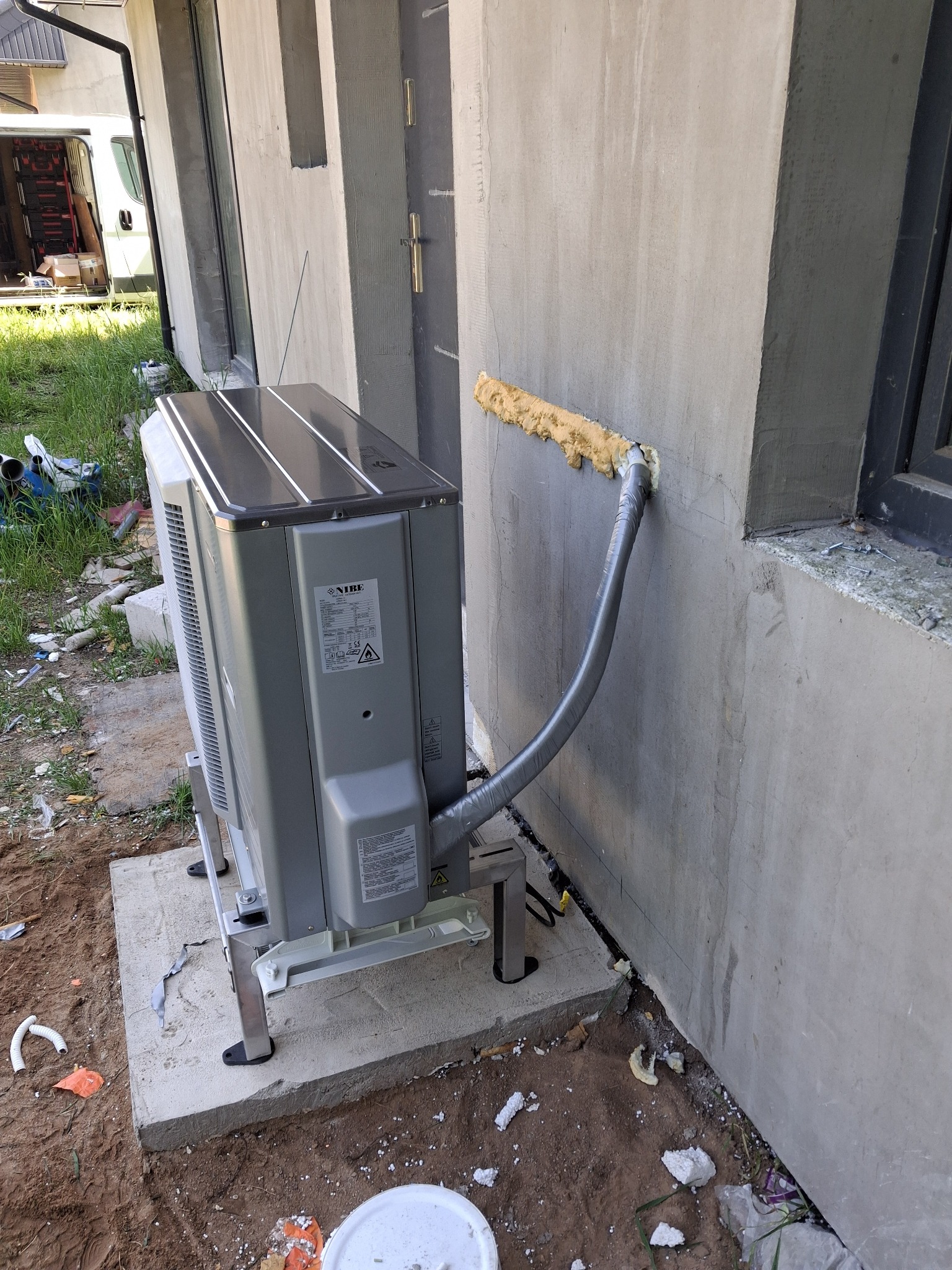 Szara jednostka zewnętrzna klimatyzacji Nibe, zamontowana na betonowej podstawie przy ścianie budynku, z widocznym przewodem zabezpieczonym pianką montażową.