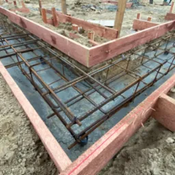 Zbrojenie fundamentów w wykopie z szalunkiem drewnianym. Widoczne pręty zbrojeniowe tworzące siatkę i betonowa podstawa. Grunt wokół wykopu.