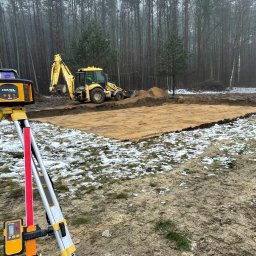 HOMEZY SPÓŁKA Z OGRANICZONĄ ODPOWIEDZIALNOŚCIĄ - Żółta niwelator laserowy na statywie, w tle wykop pod fundamenty i koparka na działce z resztkami śniegu w pochmurny dzień.