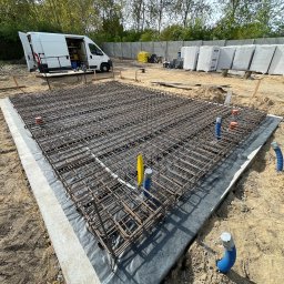 HOMEZY SPÓŁKA Z OGRANICZONĄ ODPOWIEDZIALNOŚCIĄ - Zbrojenie fundamentu na czarnej folii, widoczne rury instalacyjne, biały bus z otwartymi drzwiami i materiały budowlane w tle. Kąt ujęcia z góry.