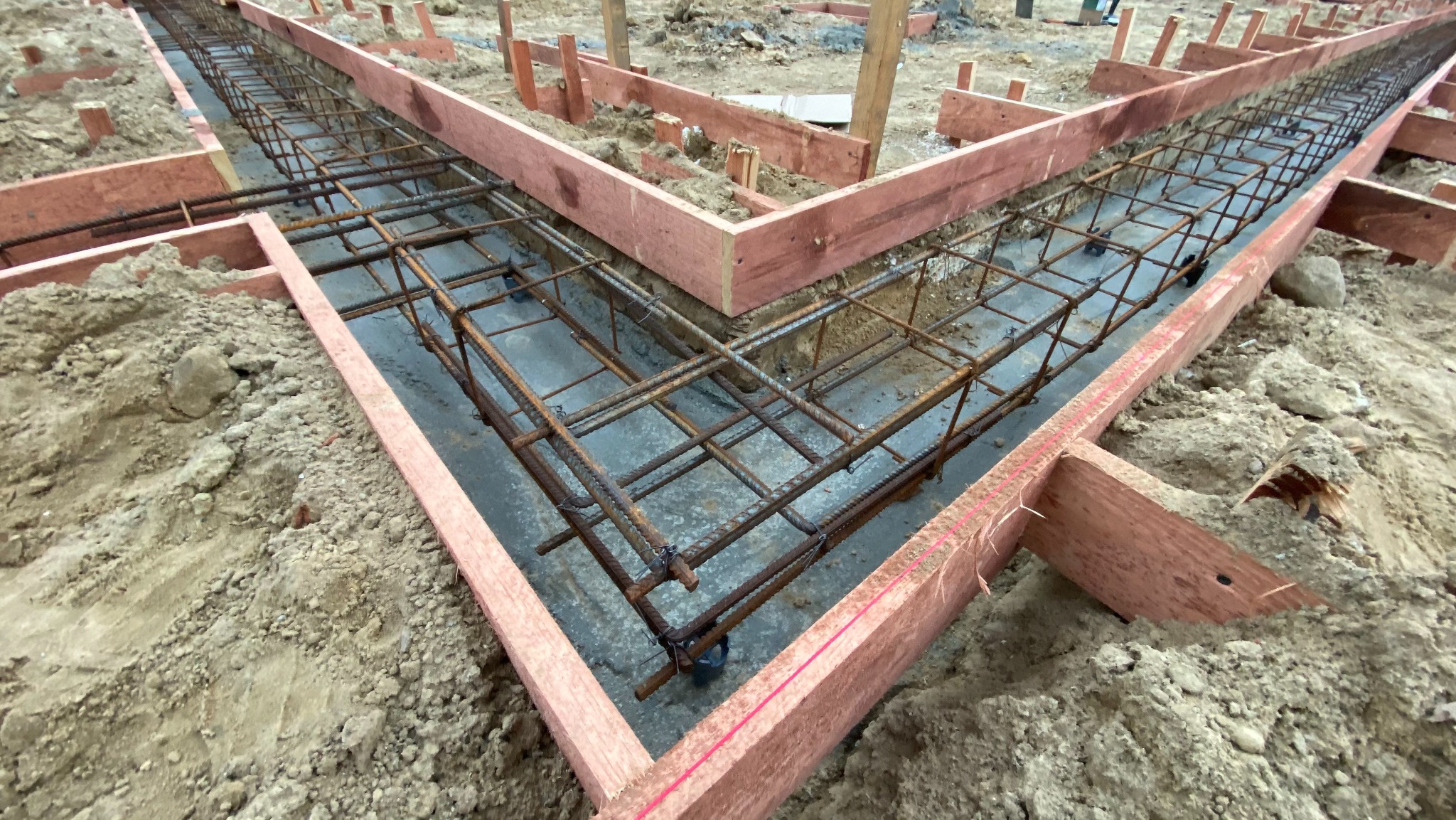 Zbrojenie fundamentów w wykopie z szalunkiem drewnianym. Widoczne pręty zbrojeniowe tworzące siatkę i betonowa podstawa. Grunt wokół wykopu.