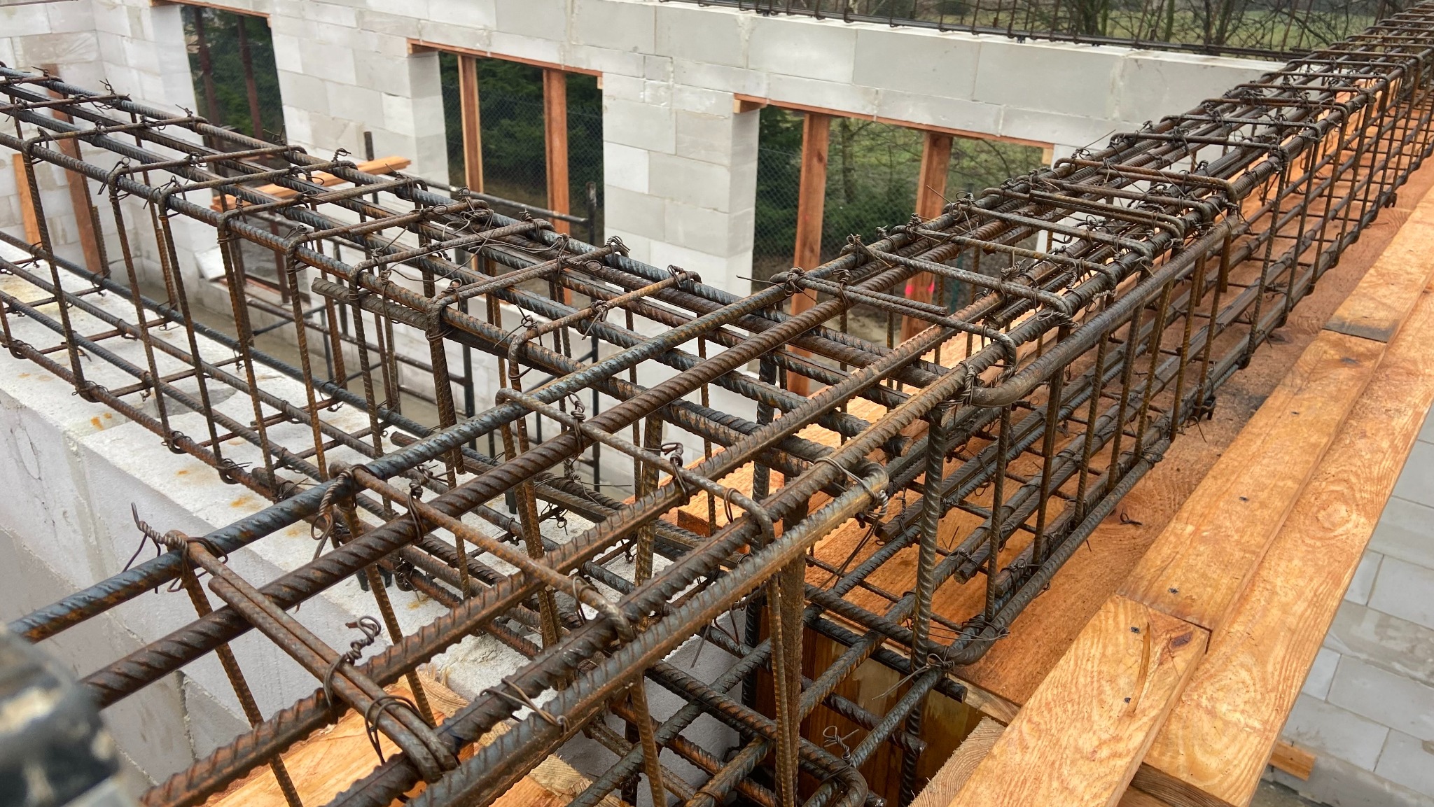 Zbrojenie stropu budynku z betonu komórkowego, widoczne pręty zbrojeniowe połączone drutem, drewniane deski szalunkowe i fragment muru z betonu komórkowego.