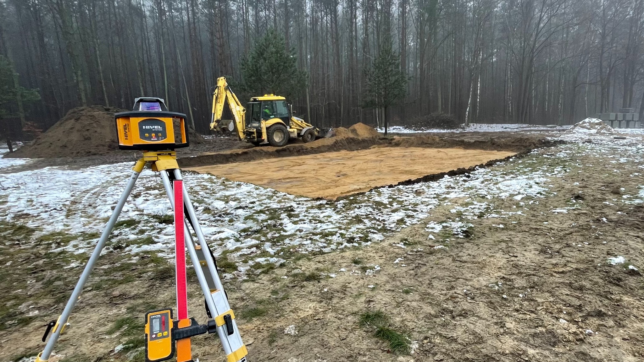 Żółta niwelator laserowy na statywie, w tle wykop pod fundamenty i koparka na działce z resztkami śniegu w pochmurny dzień.