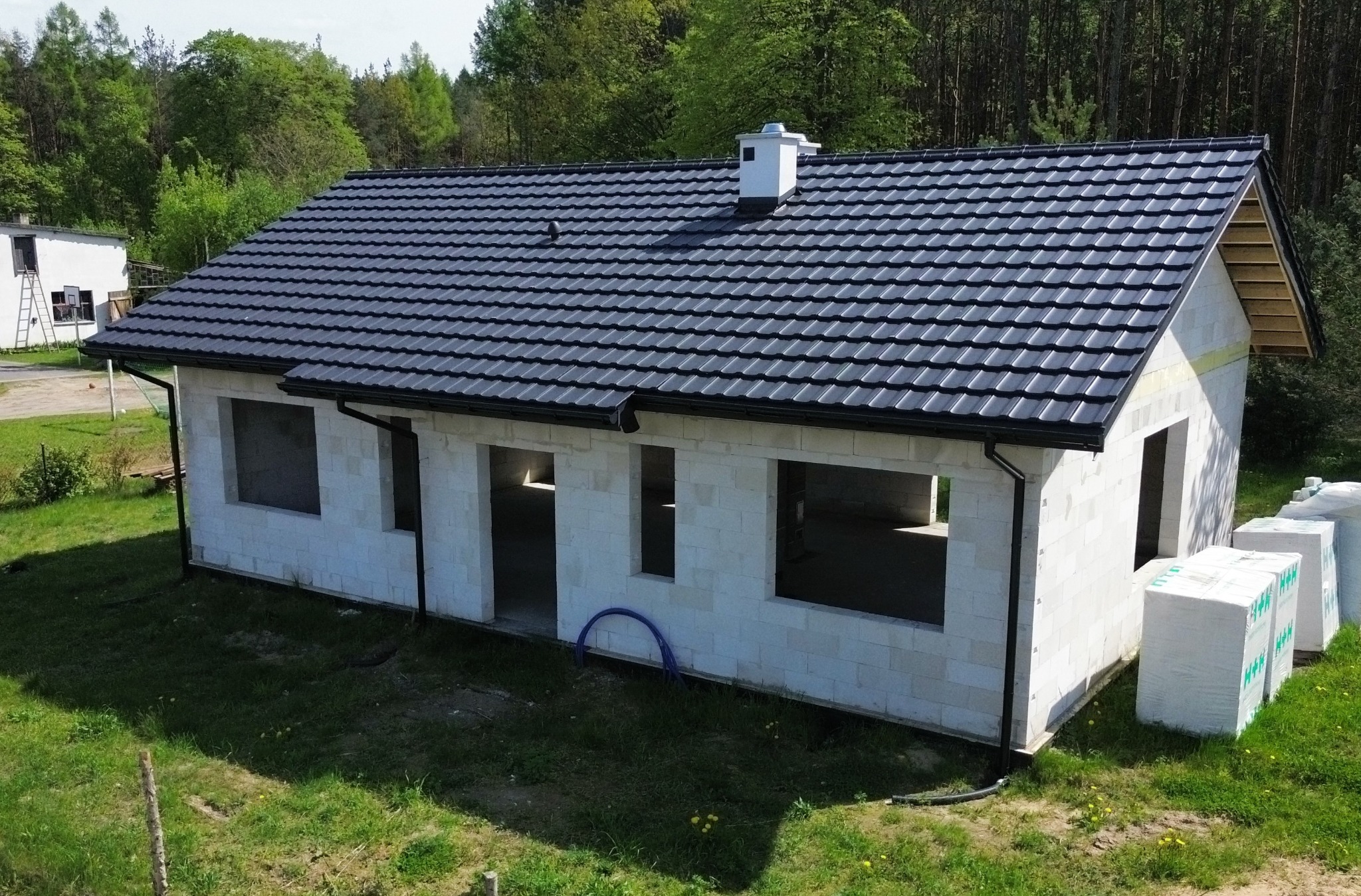 Niedokończony dom jednorodzinny z czarnym dachem z dachówki, białe ściany z pustaków, widoczne pakiety materiałów budowlanych obok budynku, w tle las.