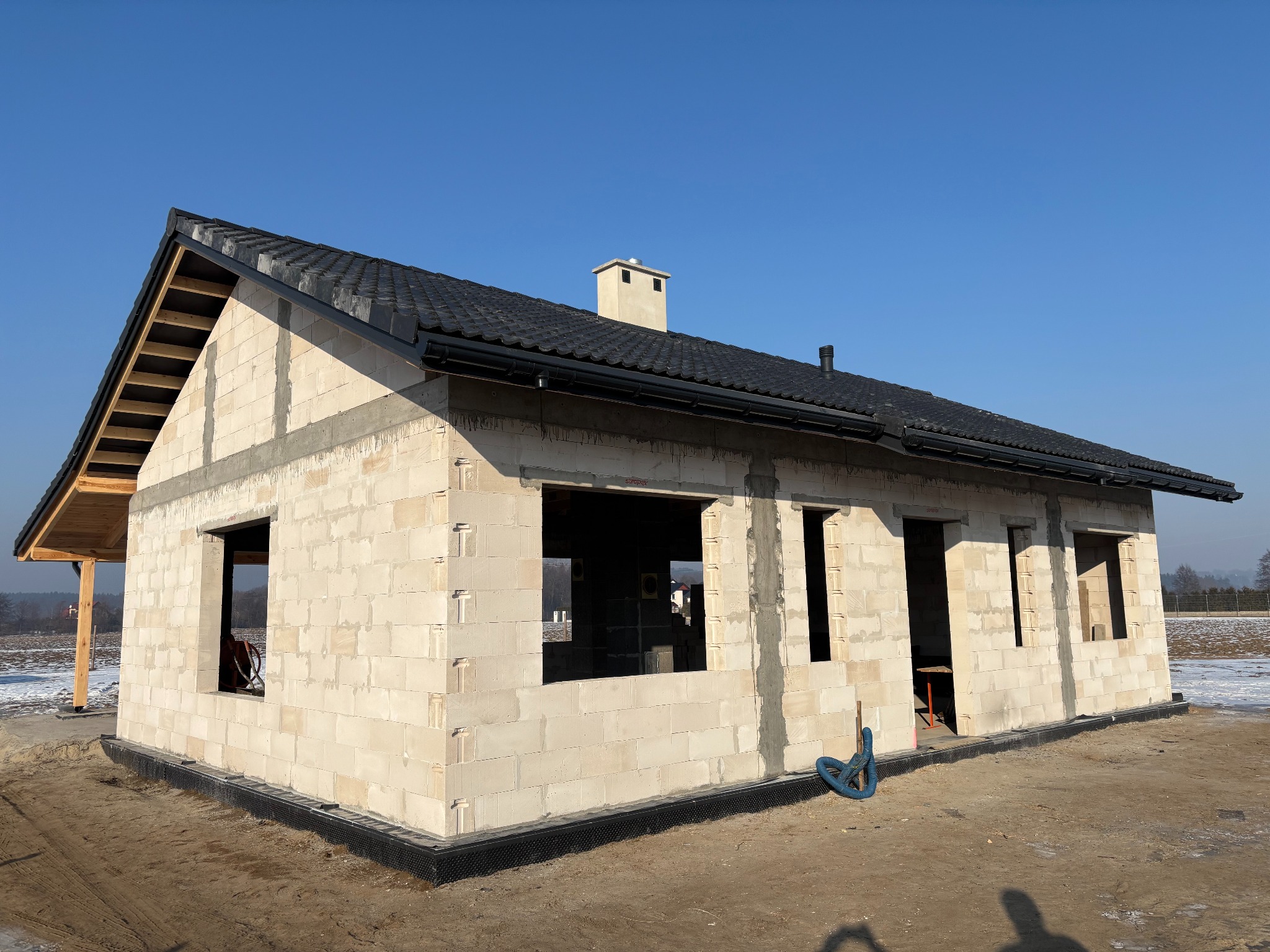 Niedokończony dom z białych bloczków, czarny dach, widoczne okna i drzwi, teren budowy, błękitne niebo. Widok z zewnątrz, wczesna faza budowy.