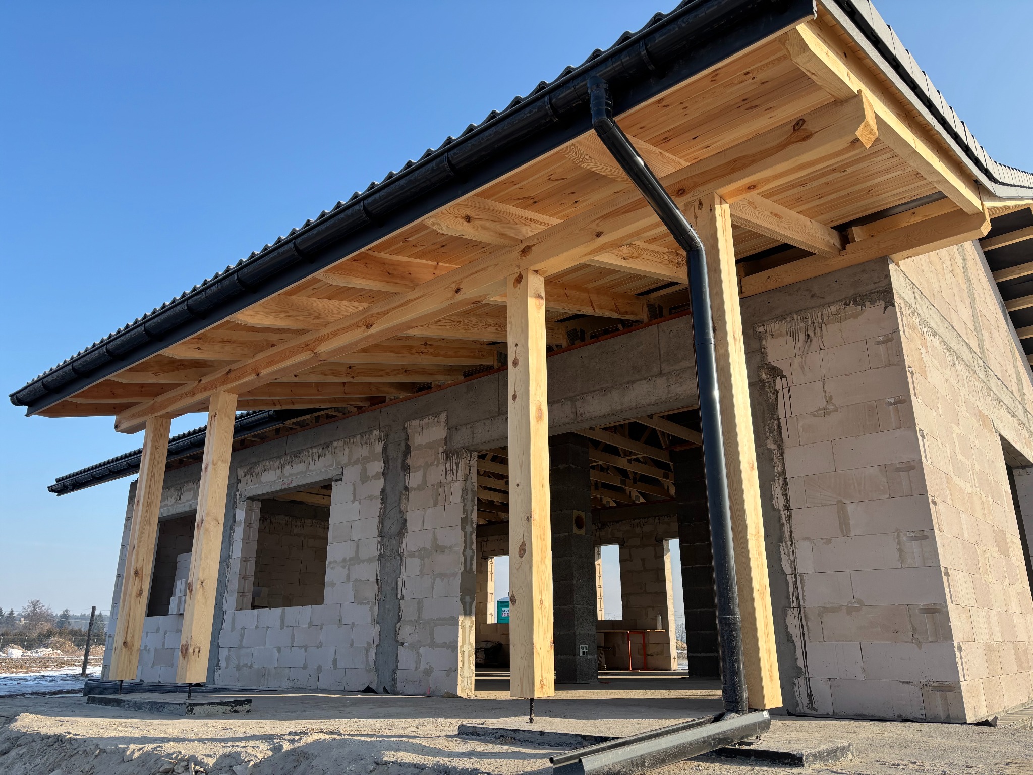 Surowy stan domu z betonu komórkowego z drewnianą konstrukcją dachu i rynnami, widok z zewnątrz w słoneczny dzień. Etap budowy z widocznymi elementami konstrukcyjnymi.