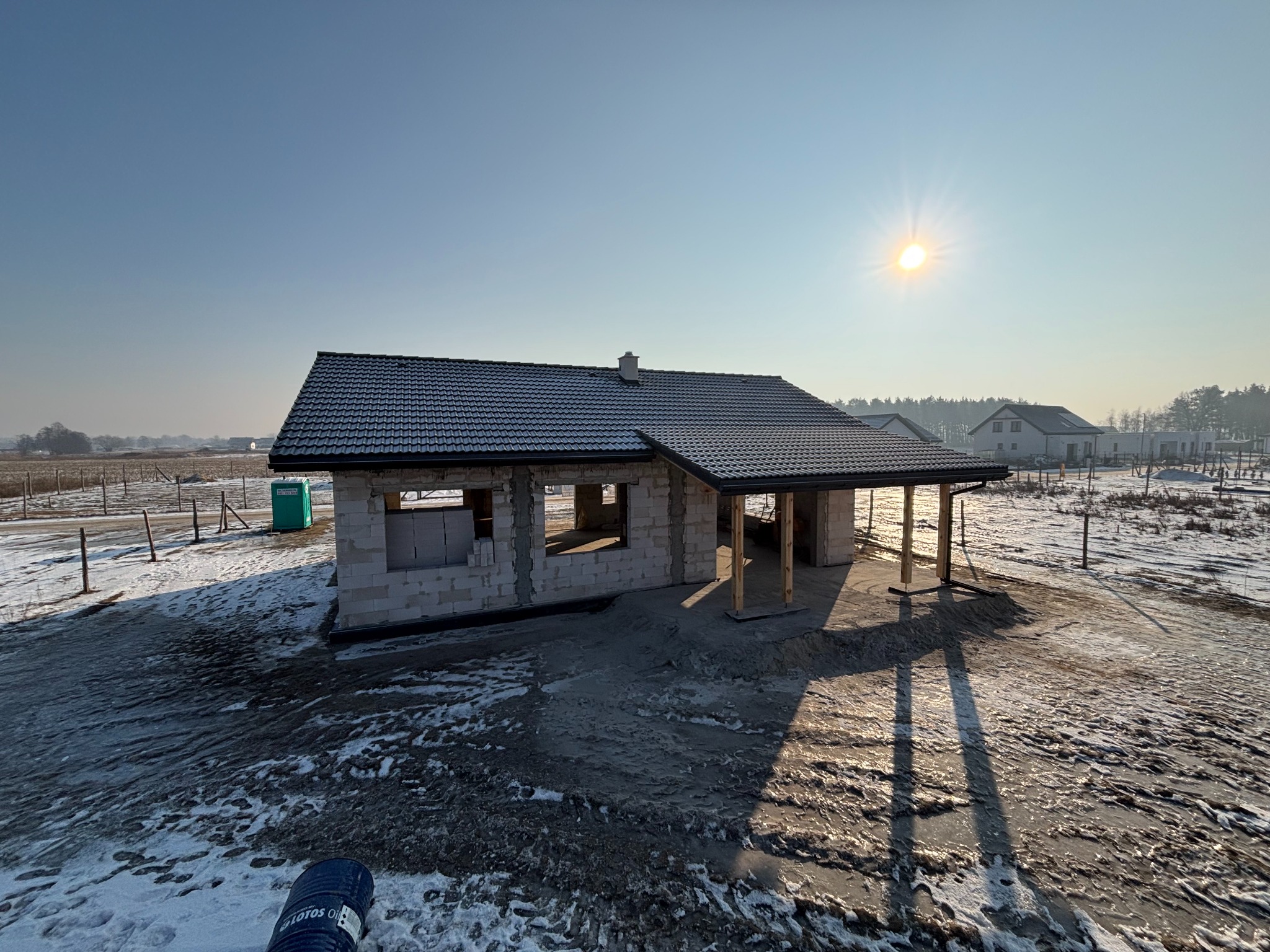 Niedokończony, parterowy dom z białego pustaka z ciemną dachówką w zimowym krajobrazie. Widoczne niezamknięte otwory okienne i drzwiowe. Słońce na błękitnym niebie.