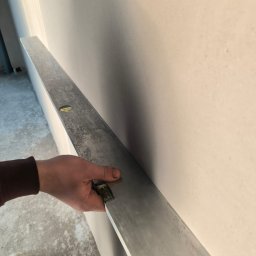 Tynki4You Andrzej Kędzia - Sprawdzanie równości ściany długą łatą aluminiową. Widoczne fragmenty podłogi i ubrania robotnika. Ściana po gładziowaniu, przygotowana do malowania.
