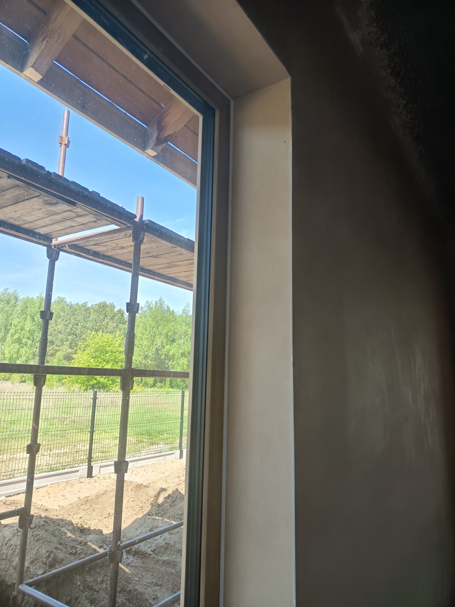 Ściana po szpachlowaniu, widok przez okno na rusztowanie i zieleń. Świeżo położona gładź na ścianie wewnątrz budynku w trakcie prac wykończeniowych.