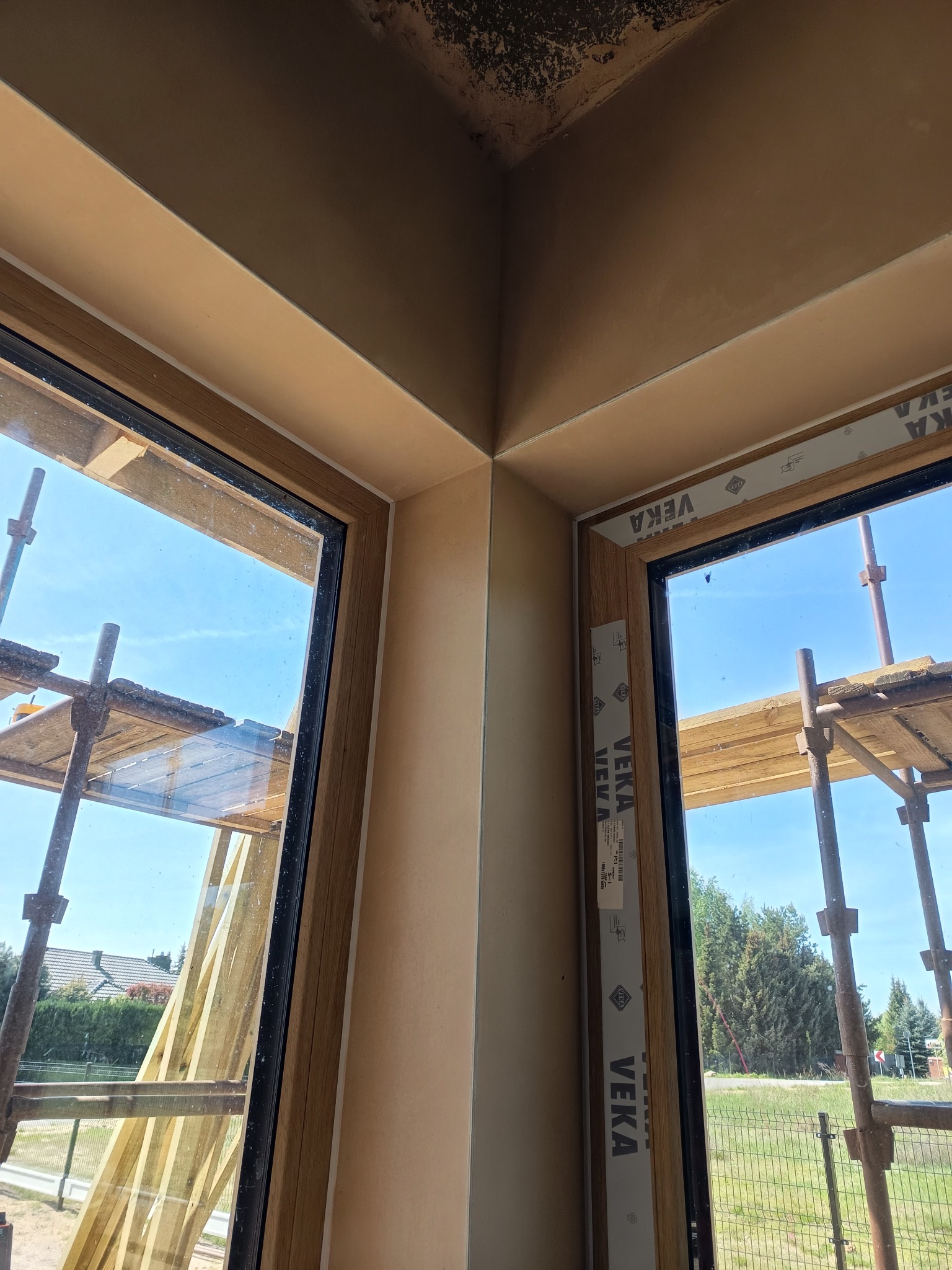 Wykończone tynki w narożniku okiennym w Żelazkowie, widok z bliska. Na zewnątrz rusztowanie i błękitne niebo. Detal wykończenia ścian.