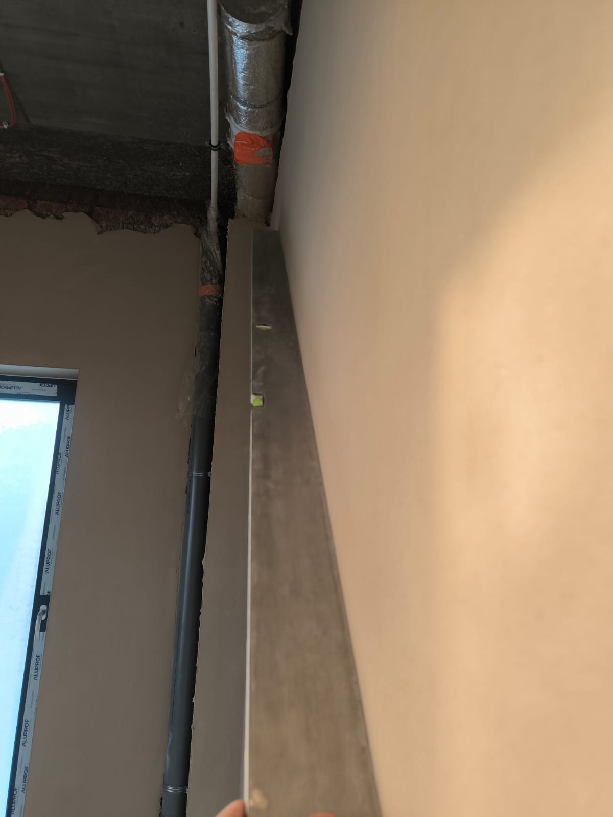 Ściana po tynkowaniu, sprawdzana poziomicą. Widoczny fragment okna i rura instalacyjna przy ścianie. Szara, gładka powierzchnia tynku.