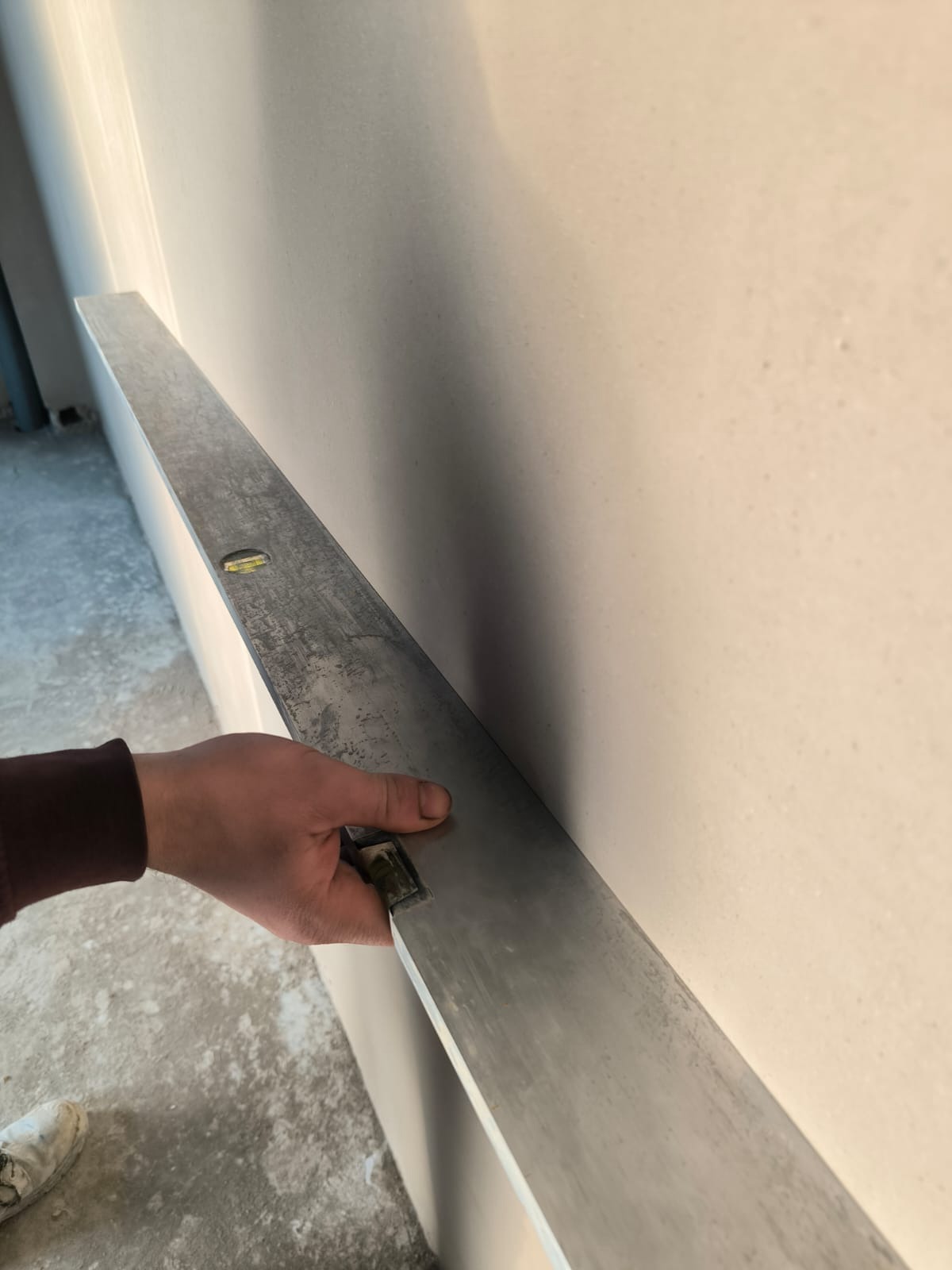 Sprawdzanie równości ściany długą łatą aluminiową. Widoczne fragmenty podłogi i ubrania robotnika. Ściana po gładziowaniu, przygotowana do malowania.