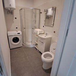 FACH EKSPERT JACEK KUSKA - Jasna łazienka z kabiną prysznicową, pralką i toaletą. Minimalistyczny design, szare płytki podłogowe, białe ściany i wyposażenie. Widok zza uchylonych drzwi.