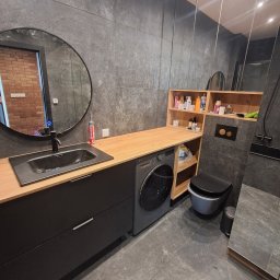 FACH EKSPERT JACEK KUSKA - Nowoczesna łazienka z szarą płytką, okrągłym lustrem i drewnianym blatem nad pralką. Uwagę zwraca minimalistyczna toaleta i zabudowa meblowa.