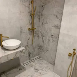 Elegancka łazienka z marmurowymi płytkami, złotą armaturą i minimalistyczną umywalką. Uwagę zwraca odpływ liniowy w podłodze.