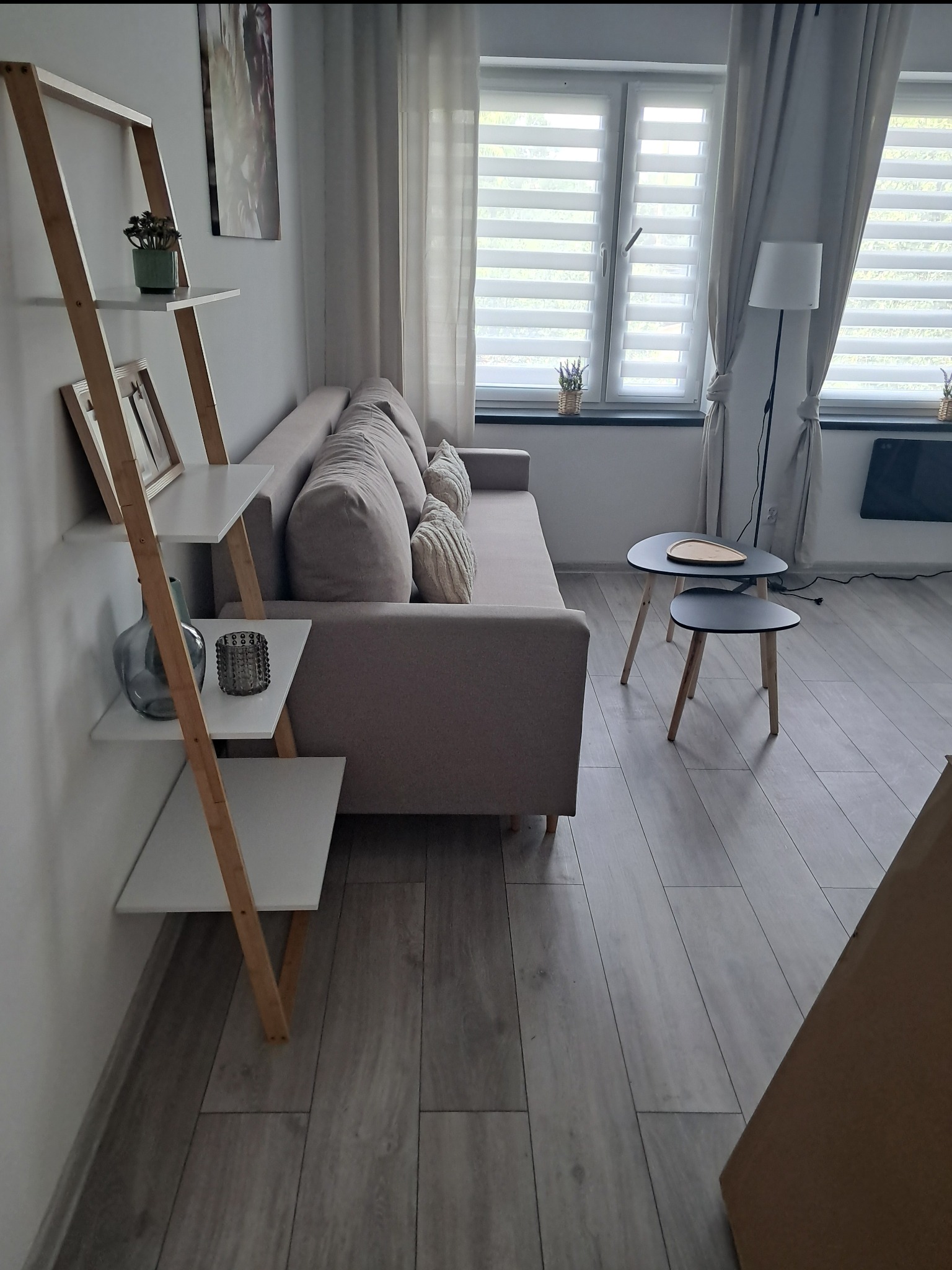 Jasny salon z drewnianą półką-drabiną, beżową sofą i stolikami kawowymi. Okno z żaluzjami i zasłonami w tle. Podłoga z paneli imitujących drewno.