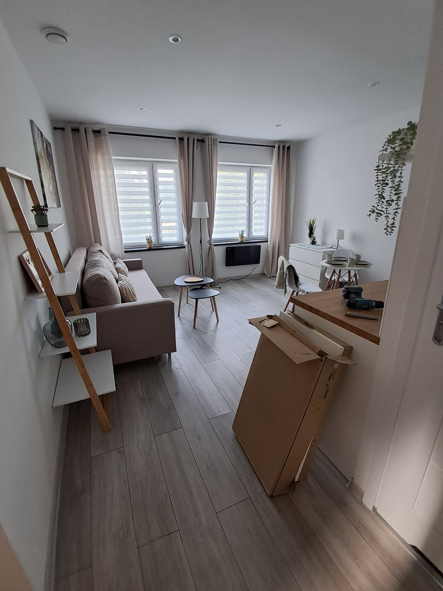 Jasne wnętrze pokoju po remoncie z drewnianą podłogą, beżową sofą i dekoracjami. Kartonowe opakowanie po meblach i narzędzia sugerują prace wykończeniowe.