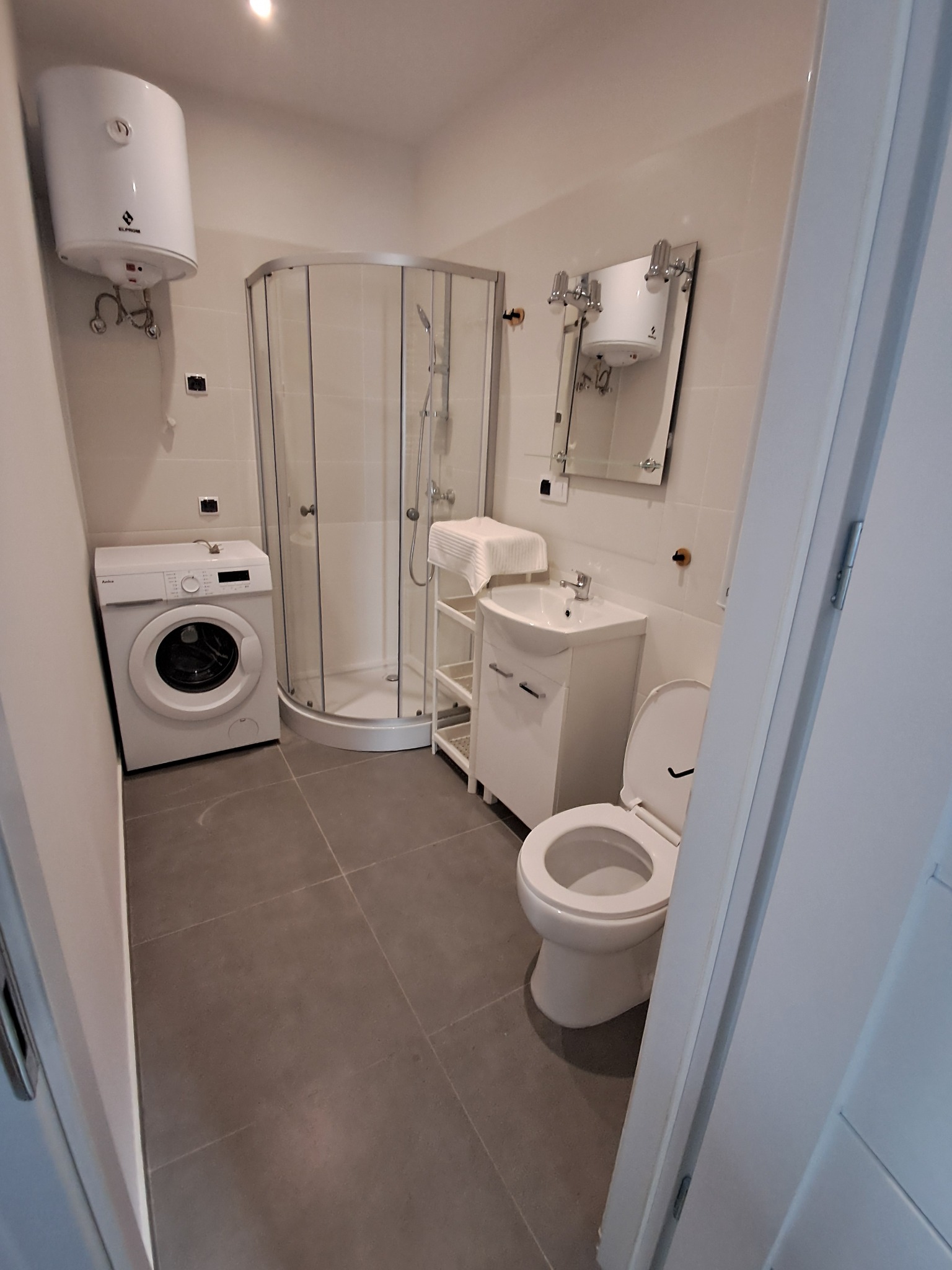 Jasna łazienka z kabiną prysznicową, pralką i toaletą. Minimalistyczny design, szare płytki podłogowe, białe ściany i wyposażenie. Widok zza uchylonych drzwi.