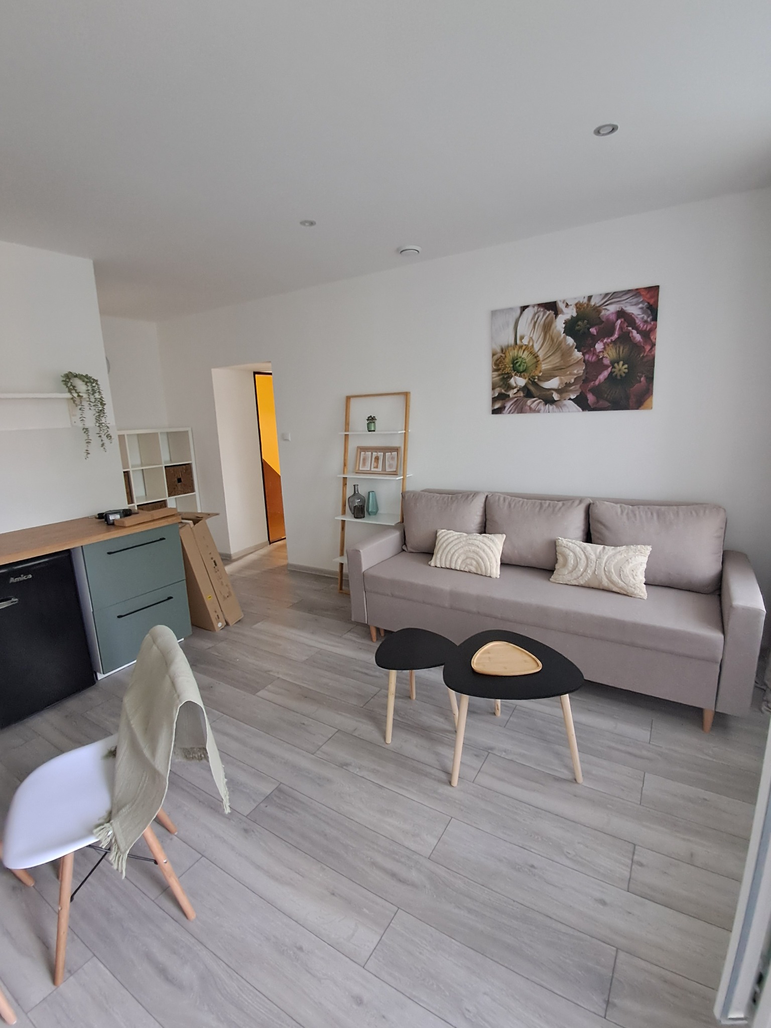 Jasne wnętrze pokoju dziennego z szarą sofą, stolikami kawowymi i drewnopodobną podłogą. W tle widoczny aneks kuchenny i regał. Całość utrzymana w nowoczesnym stylu.