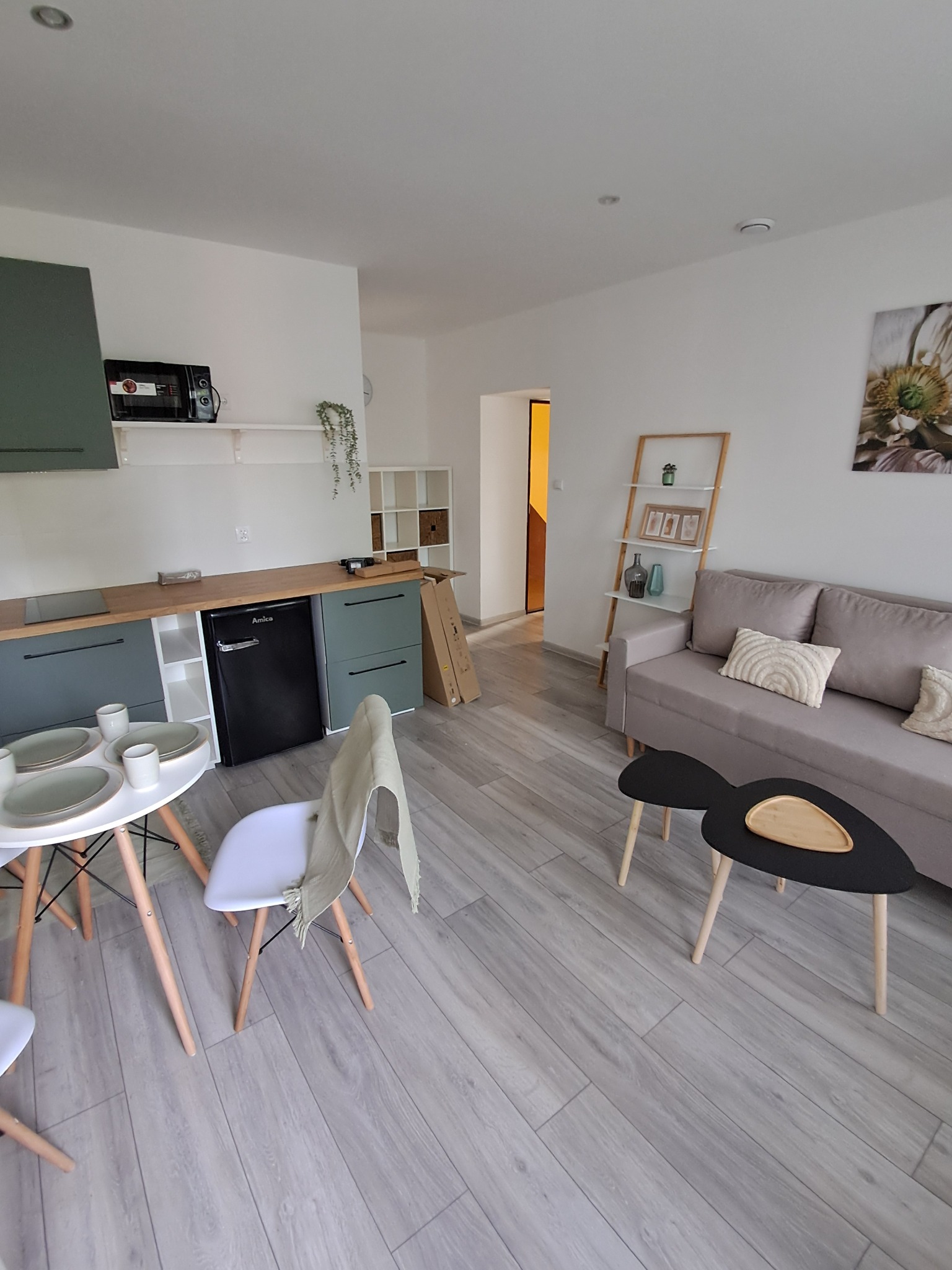 Jasne wnętrze mieszkania po remoncie: szare panele, aneks kuchenny z zielonymi szafkami, stół z zastawą, szara sofa, dekoracje. Minimalistyczny i funkcjonalny design.