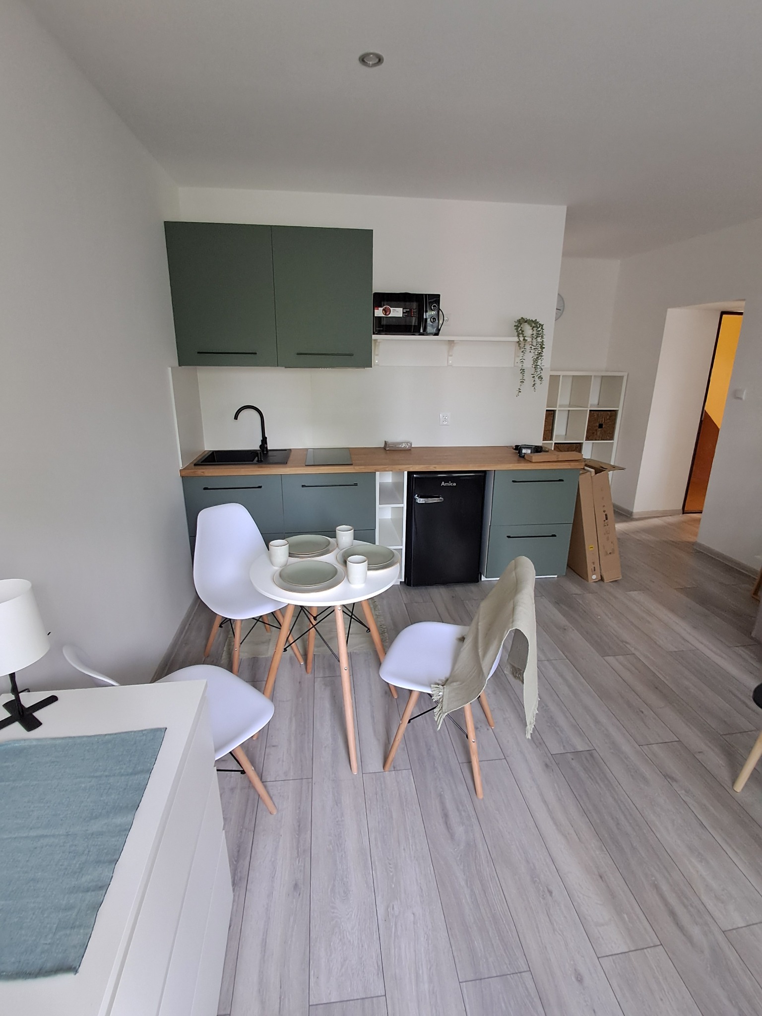 Kompletna, minimalistyczna kuchnia w odcieniach zieleni i bieli, z okrągłym stołem i krzesłami, na podłodze panele imitujące drewno, świeżo po remoncie.
