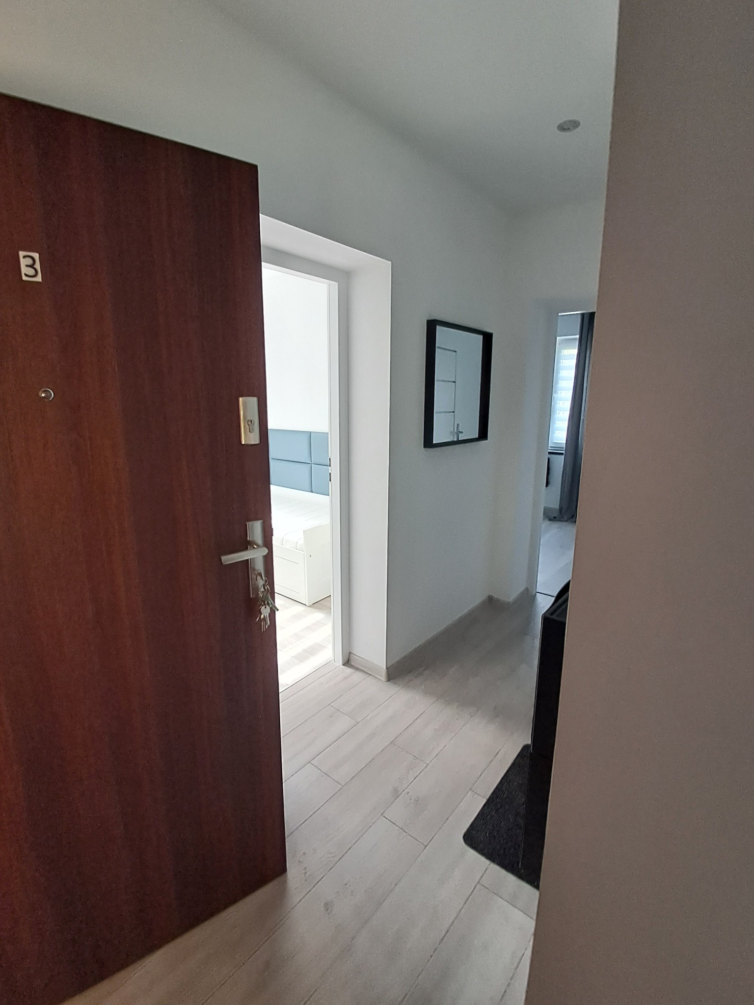 Otwarta, ciemna drewniana drzwi wejściowe z numerem '3' i kluczami, widok na jasny korytarz z lustrem i fragment sypialni w tle.