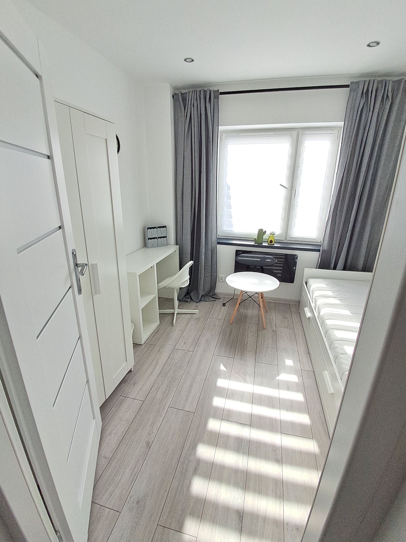 Jasne wnętrze pokoju z drewnianą podłogą, białymi meblami, biurkiem przy oknie z szarymi zasłonami i małym stolikiem. Widok zza uchylonych drzwi.