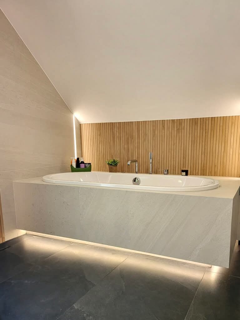 Nowoczesna wanna w obudowie z płytek imitujących kamień, podświetlona od dołu, z drewnianą ścianą w tle i akcesoriami łazienkowymi. Minimalistyczny design.