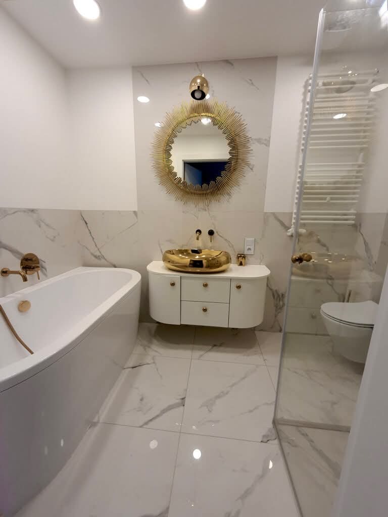Elegancka łazienka z wanną, złotą umywalką i lustrem w kształcie słońca. Białe marmurowe płytki, grzejnik drabinkowy i kabina prysznicowa tworzą luksusowy wystrój.