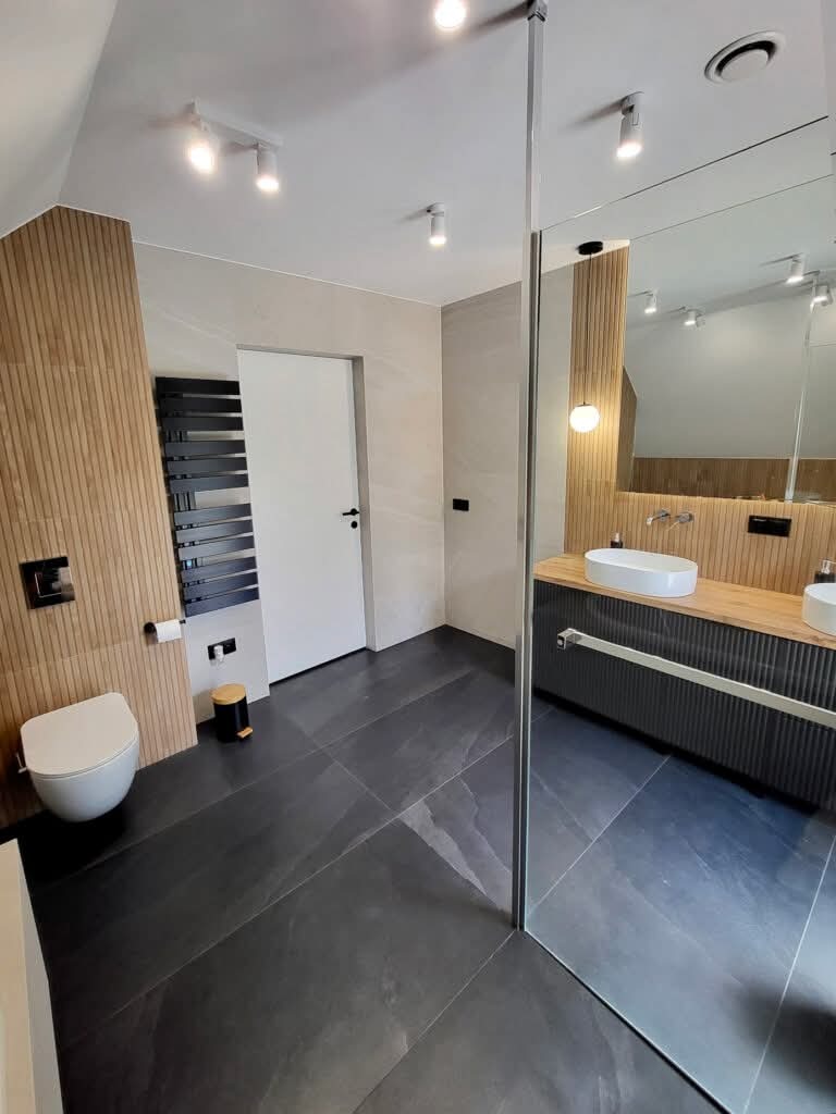 Nowoczesna łazienka z drewnianymi akcentami: płytki imitujące drewno na ścianie, ciemna podłoga, kabina prysznicowa ze szkła i minimalistyczna umywalka nablatowa.