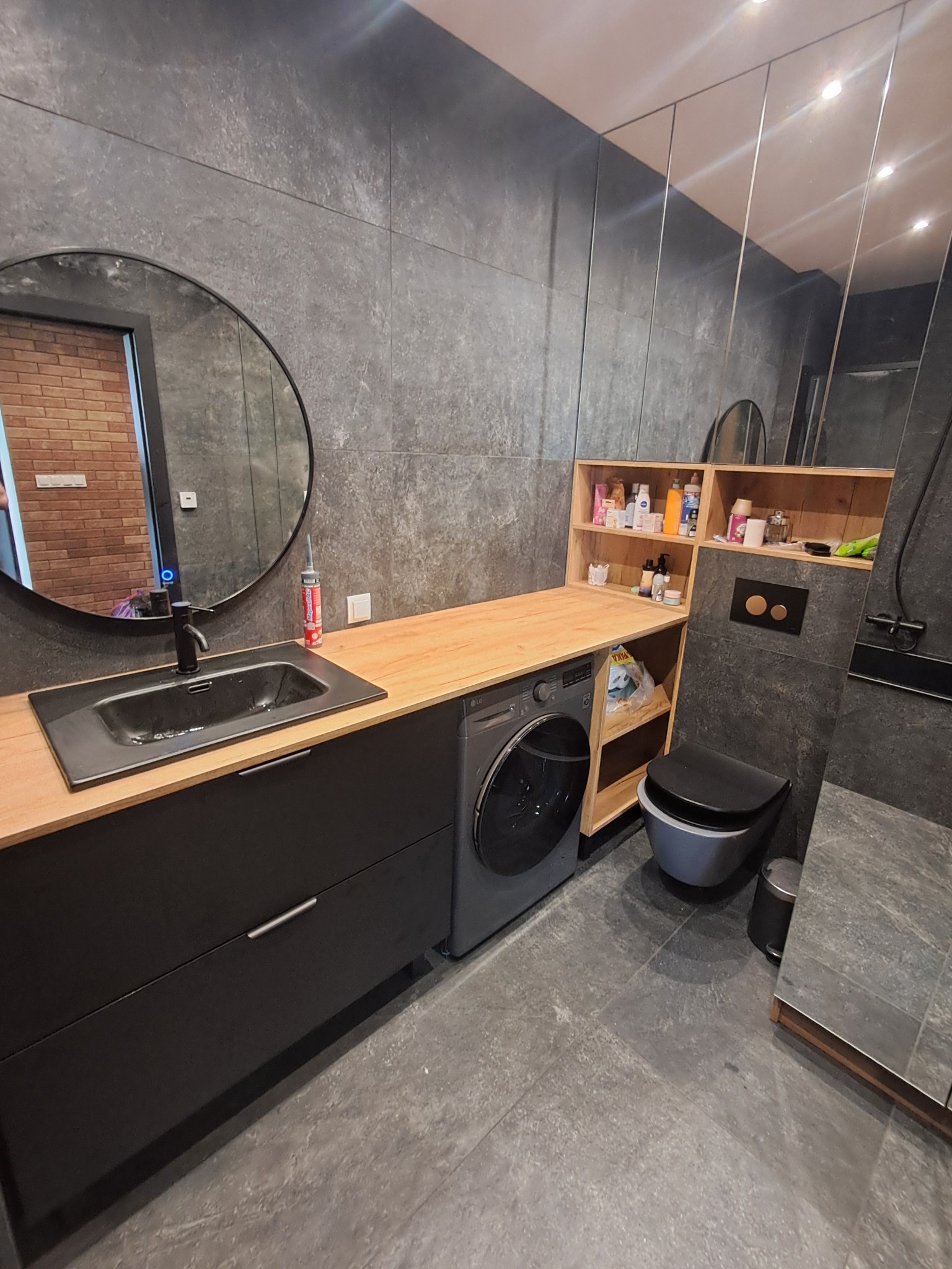 Nowoczesna łazienka z szarą płytką, okrągłym lustrem i drewnianym blatem nad pralką. Uwagę zwraca minimalistyczna toaleta i zabudowa meblowa.