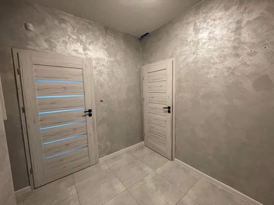 Wykończone wnętrze z dekoracyjną szarą ścianą i dwoma białymi drzwiami z poziomymi przeszkleniami. Podłoga wyłożona jasnymi płytkami.