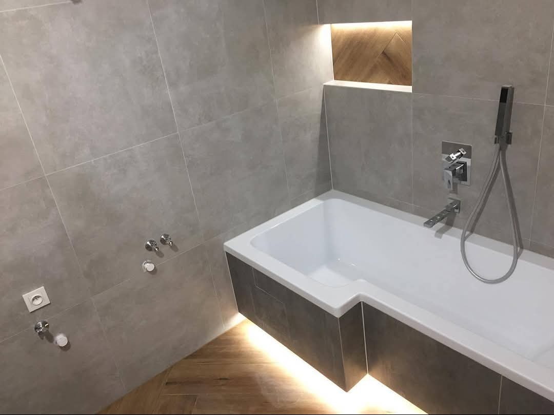 Nowoczesna łazienka z wanną obudowaną szarą mozaiką i podświetleniem LED, jasne płytki na ścianach, drewniany akcent w niszy i minimalistyczna armatura.