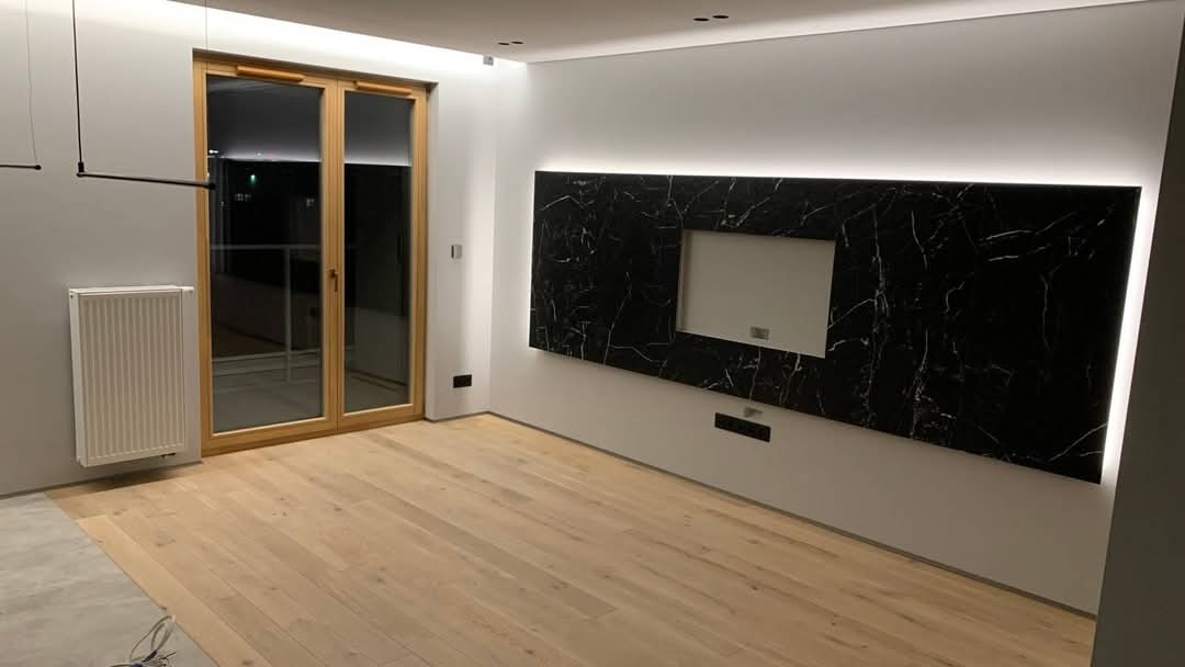 Nowoczesny salon z drewnianą podłogą, marmurową ścianą TV z podświetleniem LED i drewnianymi drzwiami balkonowymi. Grzejnik biały. Minimalistyczny design.