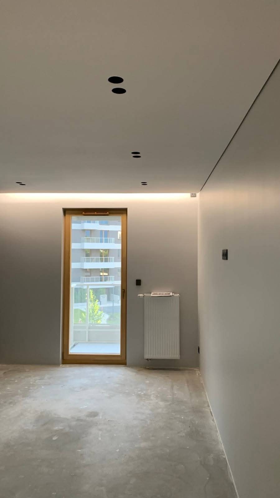 Wnętrze po gładziowaniu ścian i sufitu, widoczne punkty świetlne, okno z widokiem na budynek, grzejnik; surowy beton na podłodze. Minimalistyczny styl.