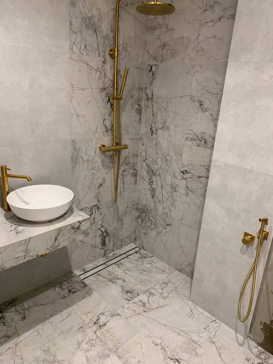 Elegancka łazienka z marmurowymi płytkami, złotą armaturą i minimalistyczną umywalką. Uwagę zwraca odpływ liniowy w podłodze.