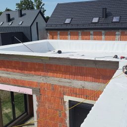 cousinroof - Nowoczesny dach płaski w trakcie hydroizolacji, widok z góry na budynek z czerwonej cegły i okoliczne domy z ciemnymi dachami. Prace dekarskie w słoneczny dzień.