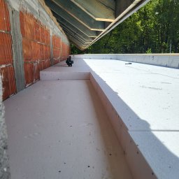 cousinroof - Ocieplenie poddasza z użyciem białych płyt styropianowych, widok z góry na fragment konstrukcji dachu i ściany z czerwonej cegły. W tle zieleń drzew.