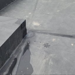 cousinroof - Szczegółowe ujęcie płaskiego dachu z papy bitumicznej, ukazujące łączenia i detale wykończenia, z widocznymi śladami wilgoci i zabrudzeń.