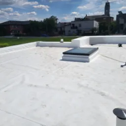 Biały dach płaski z świetlikiem i elementami wentylacyjnymi na tle zabudowań miejskich i błękitnego nieba z chmurami. Widoczne ślady na powierzchni dachu.