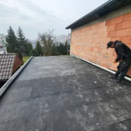 Praca na dachu płaskim: dekarz uszczelnia połączenie papy bitumicznej przy ścianie z czerwonej cegły. Widoczne ślady butów na papie i elementy rynny.