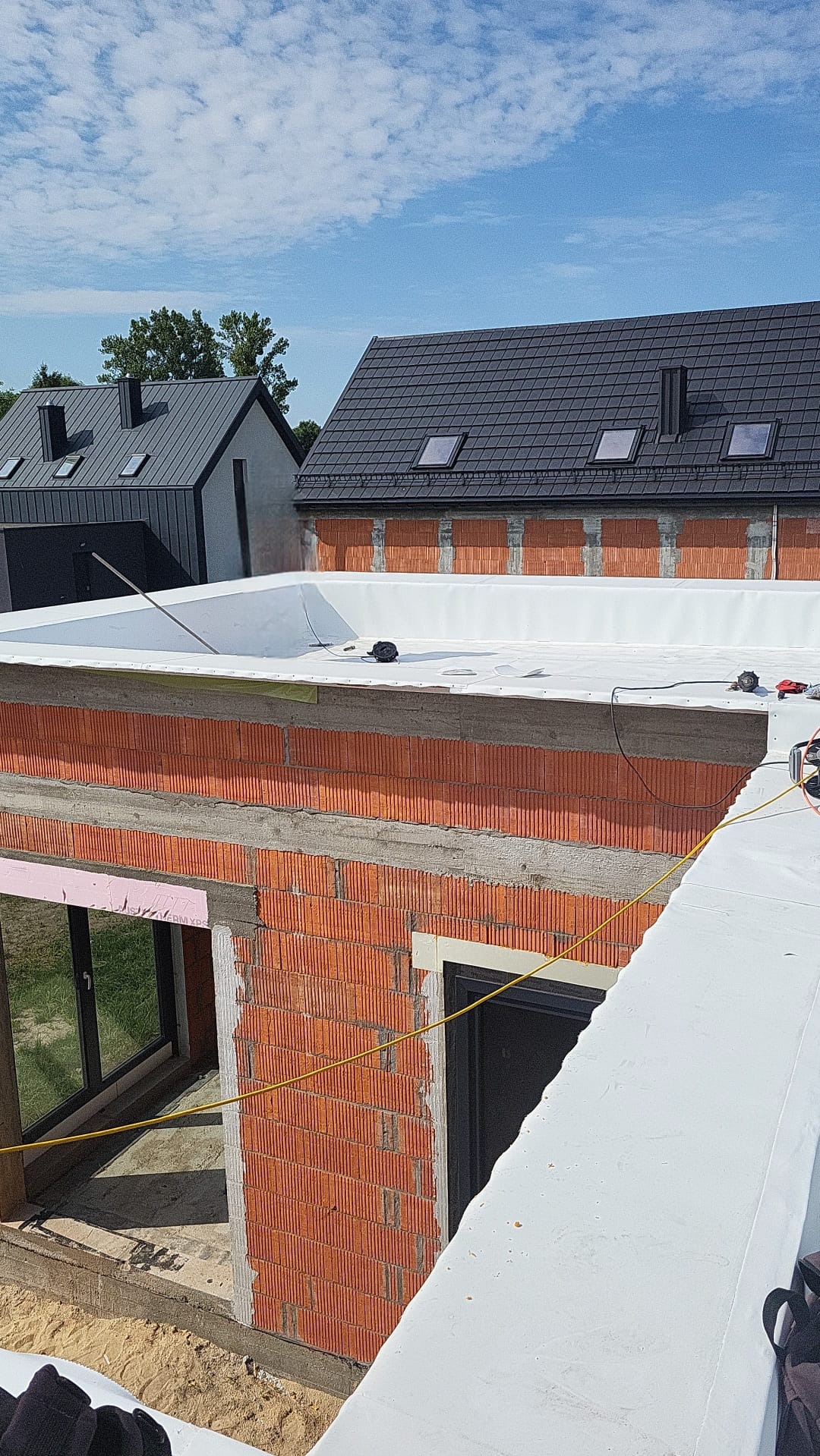 Nowoczesny dach płaski w trakcie hydroizolacji, widok z góry na budynek z czerwonej cegły i okoliczne domy z ciemnymi dachami. Prace dekarskie w słoneczny dzień.