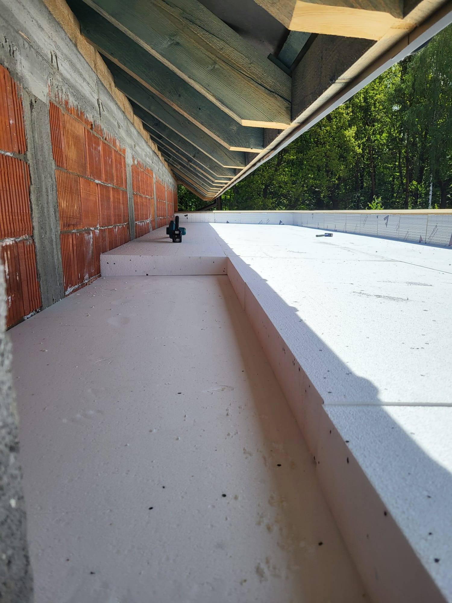 Ocieplenie poddasza z użyciem białych płyt styropianowych, widok z góry na fragment konstrukcji dachu i ściany z czerwonej cegły. W tle zieleń drzew.