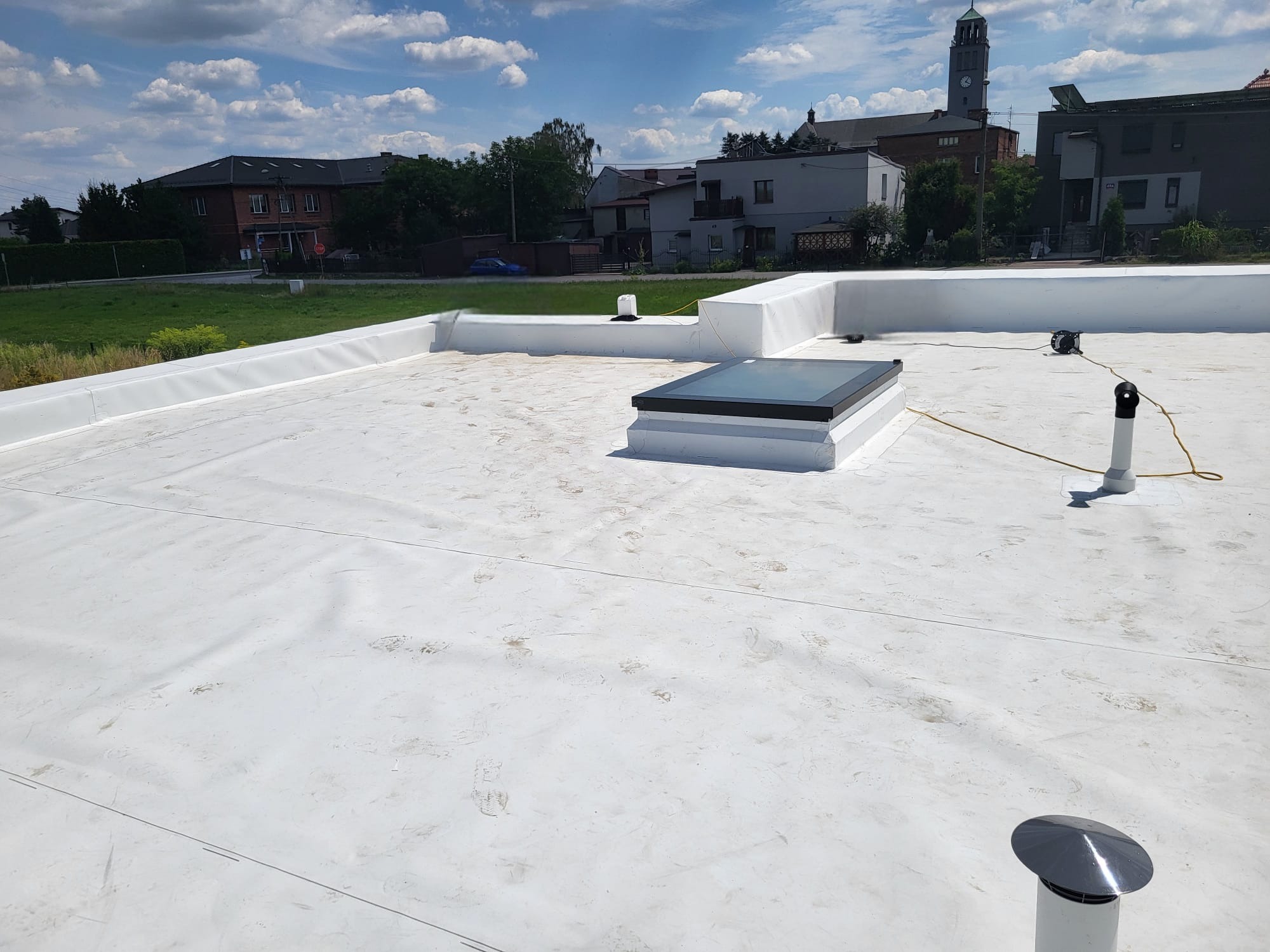Biały dach płaski z świetlikiem i elementami wentylacyjnymi na tle zabudowań miejskich i błękitnego nieba z chmurami. Widoczne ślady na powierzchni dachu.