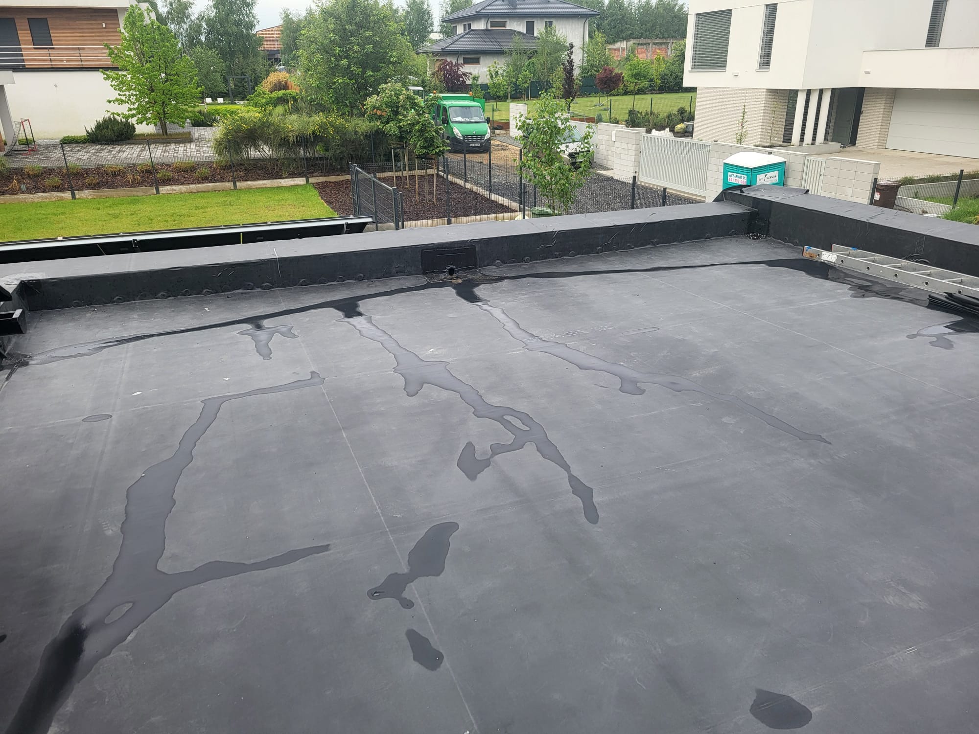 Czarna, płaska powierzchnia dachu z widocznymi śladami wody, w tle zabudowa jednorodzinna i zieleń. Widoczna drabina i elementy wykończenia dachu.