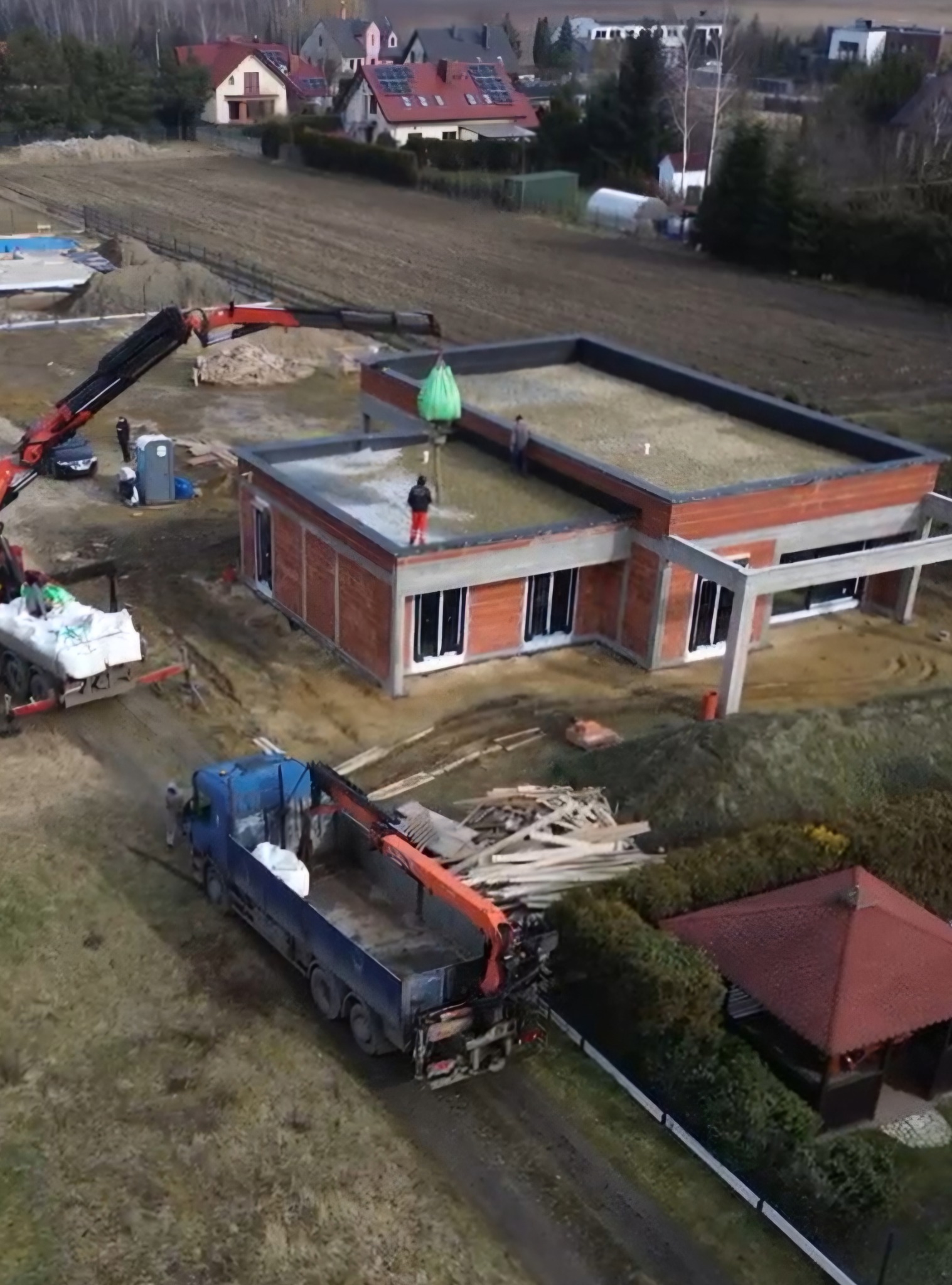 Budowa domu jednorodzinnego z płaskim dachem. Na dachu pracownicy i dźwig podający materiały. Ujęcie z drona ukazuje postęp prac na tle zabudowy i zieleni.