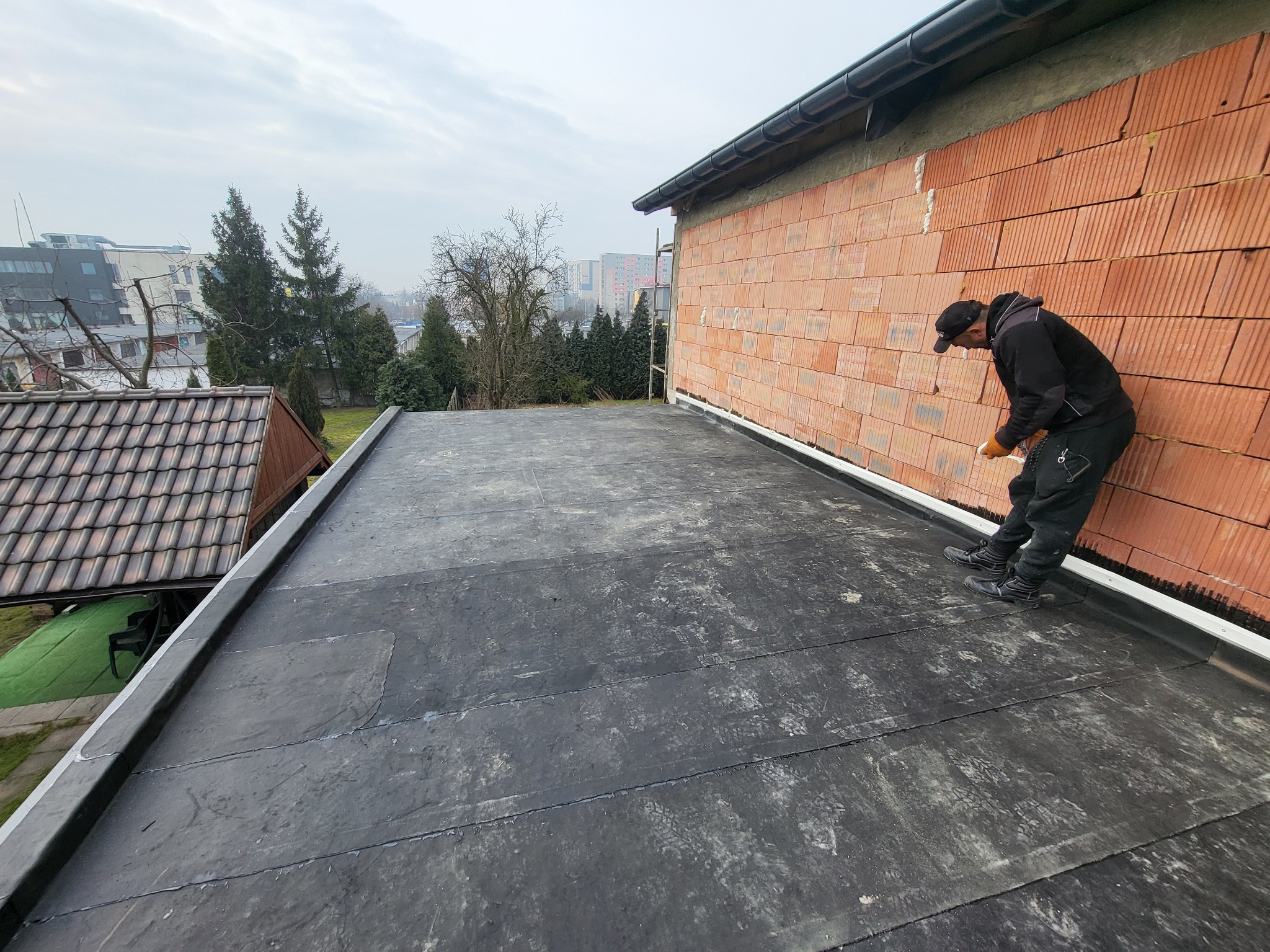 Praca na dachu płaskim: dekarz uszczelnia połączenie papy bitumicznej przy ścianie z czerwonej cegły. Widoczne ślady butów na papie i elementy rynny.