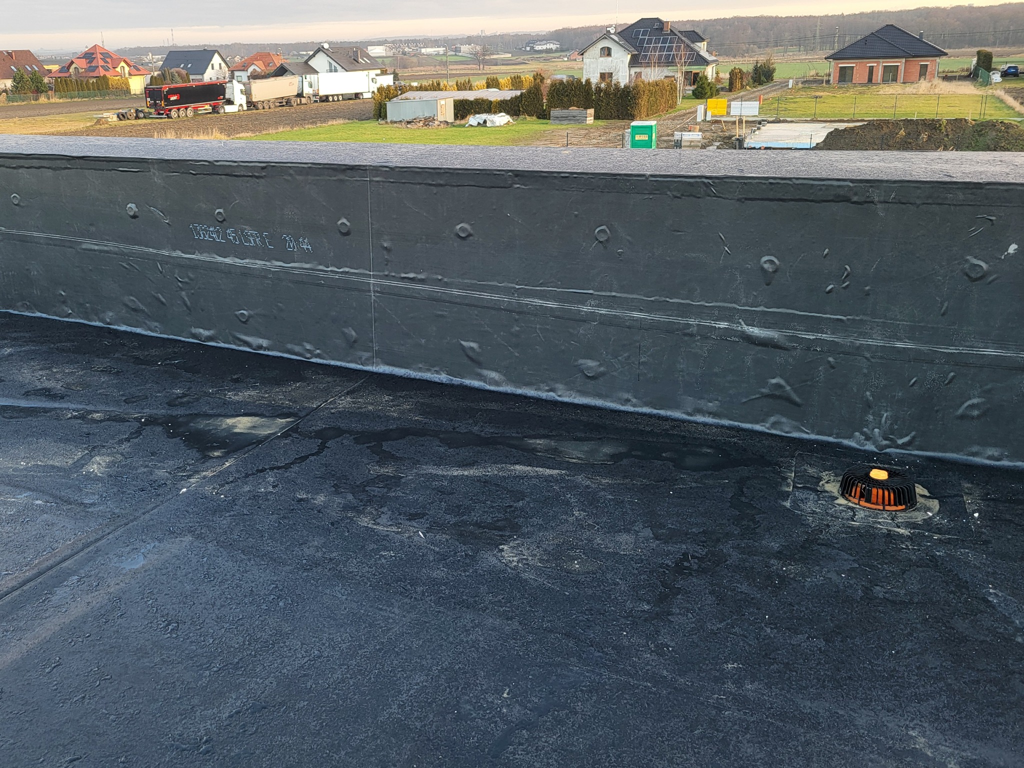 Wykończony płaski dach z czarną membraną bitumiczną, widoczny odpływ. W tle zabudowa jednorodzinna i ciężarówki na drodze, pochmurne niebo.
